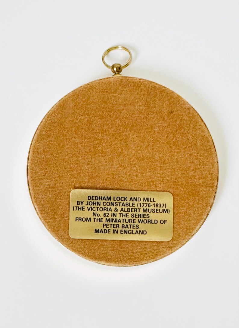 May include: A round, tan-colored miniature artwork with a gold-colored hanging loop. The artwork features a label with text that reads "DEDHAM LOCK AND MILL BY JOHN CONSTABLE (1776-1837)" and other details. Made in England.