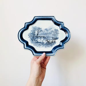 Puede incluir: Una bandeja decorativa azul y blanca con una escena de paisaje. La bandeja tiene un borde festoneado y un borde azul oscuro. El paisaje presenta árboles, un puente y una masa de agua. La bandeja está sostenida por una mano.