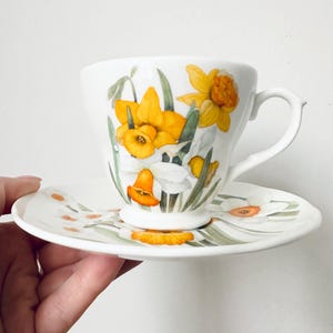 May include: White teacup and saucer set decorated with yellow and white daffodil floral design. The teacup has a curved handle and the saucer has a matching floral pattern. The set is likely made of porcelain or ceramic.