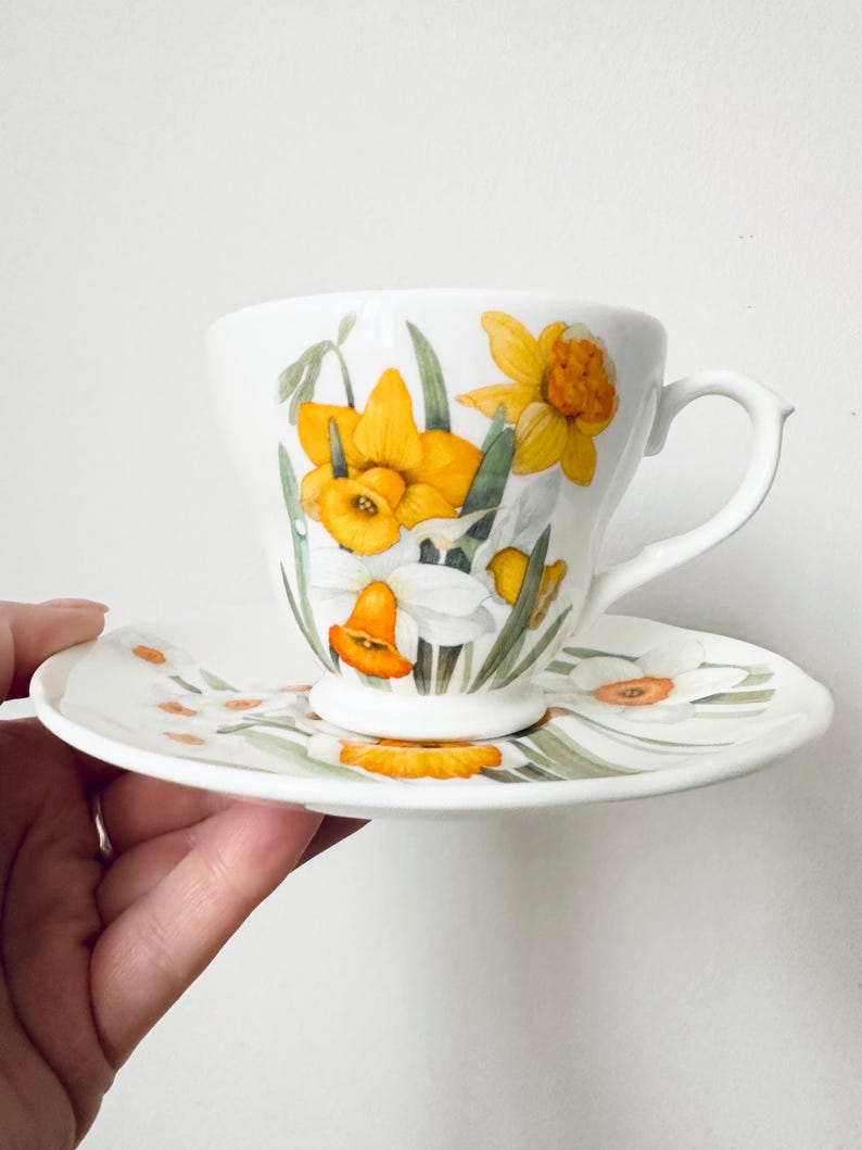 May include: White teacup and saucer set decorated with yellow and white daffodil floral design. The teacup has a curved handle and the saucer has a matching floral pattern. The set is likely made of porcelain or ceramic.