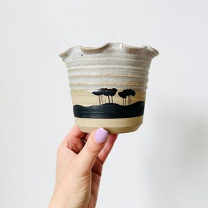 May include: A handmade ceramic planter with a frilled rim. The planter has a beige base, a black band with a landscape design, and a light grey upper section. The design includes black tree silhouettes.