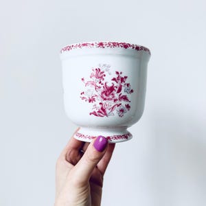 May include: White ceramic planter with a pedestal base and a floral design in shades of red. The rim and the body of the planter are decorated with a repeating floral pattern. The planter is held up by a hand with purple nail polish.