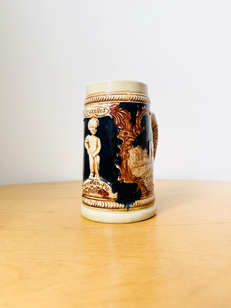 Op de afbeelding: Een klein, keramisch bierglas met een blauwe achtergrond en bruine en witte details. Het glas toont een afbeelding van een gebouw en een figuur, samen met de tekst "Bruxelles" en "Manneken-Pis".