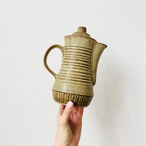 Peut inclure: Une théière en céramique beige avec un couvercle, une anse et un bec verseur. La théière présente des rayures horizontales marron et une surface texturée. La théière est tenue par une main sur fond blanc.