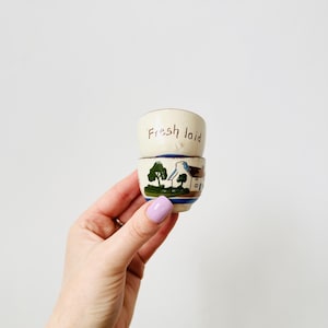 Puede incluir: Dos pequeños hueveras de cerámica color crema. La taza superior tiene las palabras "Fresh laid" en escritura marrón. La taza inferior presenta una escena pintada de una casa, árboles y una franja azul y marrón. Las tazas se sostienen en una mano.