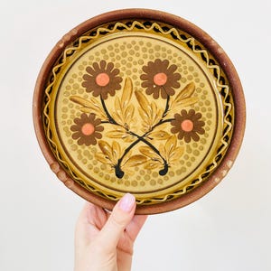 May include: Round ceramic plate with a floral design. The plate features brown flowers with orange centers and brown leaves on a yellow background. The rim is brown with a decorative edge.