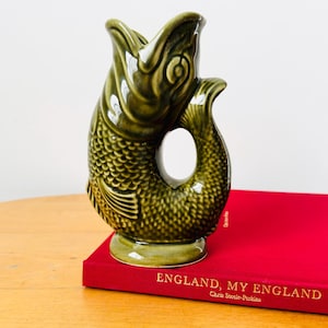 May include: A glossy olive green ceramic fish-shaped pitcher with a textured scale pattern and an open mouth spout. The pitcher rests on a red book titled "ENGLAND, MY ENGLAND" by Chris Steele-Perkins, featuring a curved handle formed by the tail.