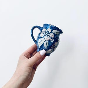 Op de afbeelding: Een kleine keramieken melkkannetje met een diepblauwe glazuurlaag, voorzien van handgeschilderde witte bloemmotieven en groene bladaccenten. Het kannetje heeft een gebogen handvat, een kleine schenktuit en een textuur met zichtbare penseelstreken.