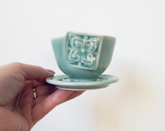 May include: A small, light teal ceramic teacup and saucer set. The teacup features a floral design on one side. The set is held in a hand against a white background. The teacup and saucer are both round.
