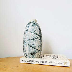 Puede incluir: Un jarrón de cerámica blanco con un patrón geométrico azul y blanco. El jarrón está sobre un libro titulado "Mad About The House" de Kate Watson-Smyth.