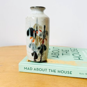May include: A small, cylindrical ceramic vase with a speckled off-white glaze. The exterior features a hand-painted botanical motif in shades of black, muted green, and bright orange. The vase has a narrow neck and a dark, textured rim.