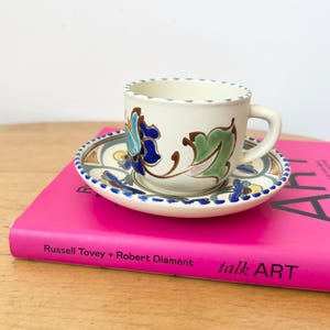 May include: A cream-colored ceramic teacup and saucer set featuring hand-painted floral motifs in shades of blue, green, and brown. The rim of both the cup and saucer is decorated with a repeating pattern of small blue dots. The set rests on a book.
