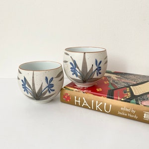 May include: Two white ceramic teacups with blue and gray floral designs, sitting on top of a gold book titled "HAIKU" with the text "edited by Jackie Hardy".