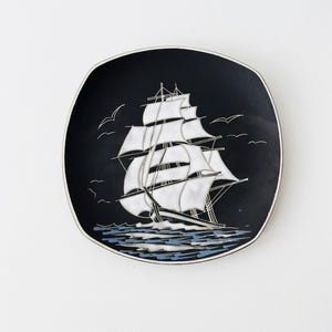 May include: A square-shaped decorative plate with rounded corners featuring a black background. A white sailing ship with multiple masts and sails is centred on the plate, with stylised blue and white waves below and white birds in the sky.