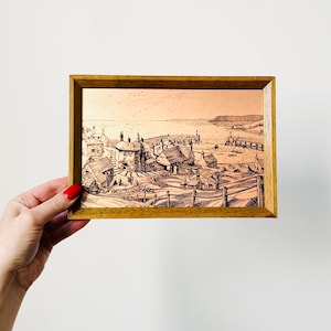 Op de afbeelding: Een ingelijst kopergravure van een kustdorp met huizen en een haven. De gravure is in zwart-wit en heeft een warme, roodbruine achtergrond.