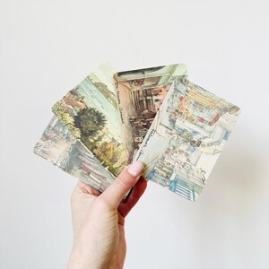 Peut inclure: Trois cartes postales vintage, tenues par une main, présentent des illustrations à l'aquarelle de scènes côtières. Les cartes postales sont rectangulaires avec des coins arrondis. Le texte "St. Mawes, Cornwall" est visible.