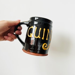 May include: A black ceramic mug with the word "Guinness" in gold lettering. The mug has a handle and a brown base. The mug is held by a person.