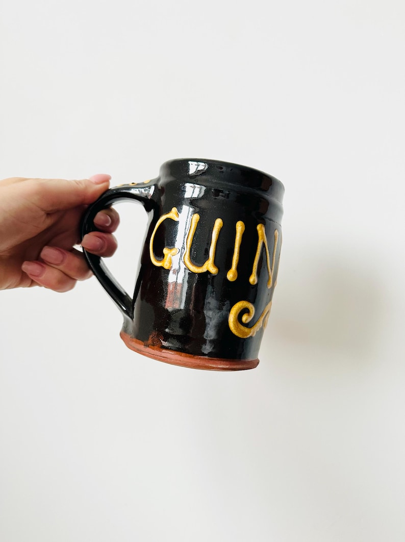May include: A black ceramic mug with the word "Guinness" in gold lettering. The mug has a handle and a brown base. The mug is held by a person.
