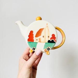 Puede incluir: Una tetera de cerámica con un diseño caprichoso que presenta una casa, árboles y flores. La tetera tiene un cuerpo blanco, un asa amarilla y un pomo amarillo en la tapa. El diseño incluye detalles en naranja, rojo, verde y negro.