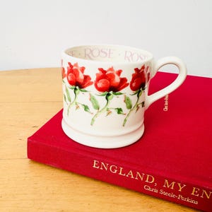 May include: A white ceramic mug featuring a repeating pattern of red roses with green stems and leaves around the exterior. The interior rim of the mug displays the word ROSE printed in a repeating sequence in a light pink font.