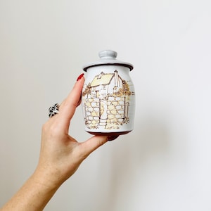 May include: White ceramic jar with a brown and yellow illustration of a house with a brick wall and a thatched roof. The jar has a lid.