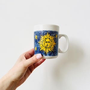 Puede incluir: Taza de cerámica blanca con un diseño de sol amarillo sobre un fondo azul con estrellas amarillas. La taza tiene un asa y un borde blancos. La taza está sujeta por una mano.