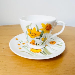 May include: White teacup and saucer set decorated with yellow and white daffodil floral pattern. The teacup has a curved handle and sits on a matching saucer. The design includes green leaves and stems.
