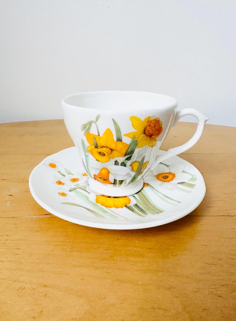 May include: White teacup and saucer set decorated with yellow and white daffodil floral pattern. The teacup has a curved handle and sits on a matching saucer. The design includes green leaves and stems.
