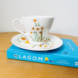 May include: White teacup and saucer set decorated with watercolor daffodils. The teacup sits on a matching saucer, both adorned with yellow and white floral designs. The set rests on a blue book with the word "LAGOM" on the cover.