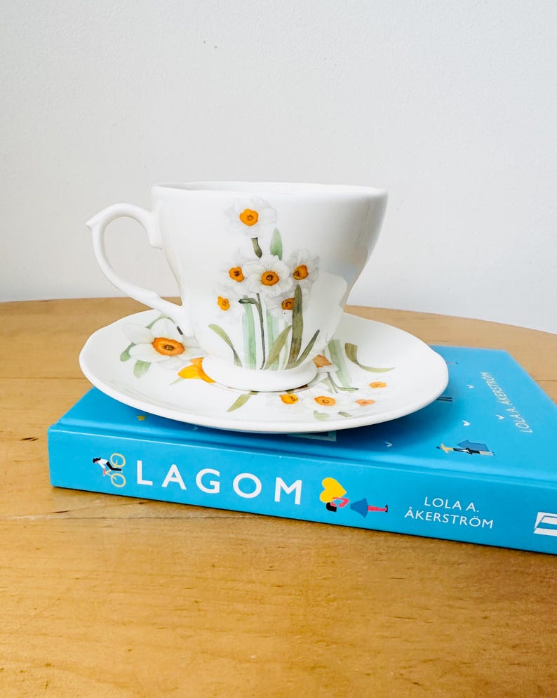 May include: White teacup and saucer set decorated with watercolor daffodils. The teacup sits on a matching saucer, both adorned with yellow and white floral designs. The set rests on a blue book with the word "LAGOM" on the cover.