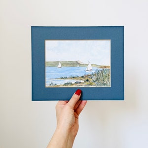 Puede incluir: Una pintura de acuarela enmarcada de un paisaje mar&iacute;timo con tres veleros en la distancia. La pintura est&aacute; ambientada en un cielo azul con nubes blancas. El agua es de color azul claro y la costa es de color verde. La pintura est&aacute; enmarcada en un marco azul oscuro.