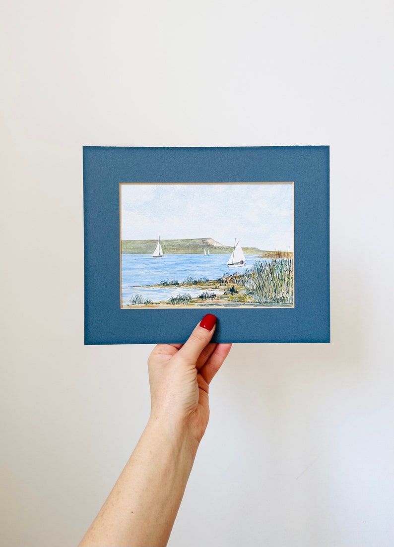 Puede incluir: Una pintura de acuarela enmarcada de un paisaje mar&iacute;timo con tres veleros en la distancia. La pintura est&aacute; ambientada en un cielo azul con nubes blancas. El agua es de color azul claro y la costa es de color verde. La pintura est&aacute; enmarcada en un marco azul oscuro.