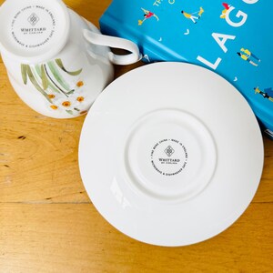 May include: White fine bone china teacup and saucer set with floral design. The teacup is upside down, revealing the floral pattern and the brand "WHITTARD OF CHELSEA". A blue book with the title "THE SWEDISH SECRET OF LIVING WELL" is in the background.
