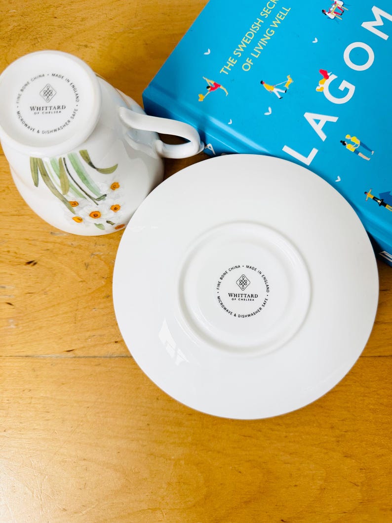 May include: White fine bone china teacup and saucer set with floral design. The teacup is upside down, revealing the floral pattern and the brand "WHITTARD OF CHELSEA". A blue book with the title "THE SWEDISH SECRET OF LIVING WELL" is in the background.