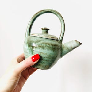 Pode incluir: Pequena chaleira de cerâmica artesanal em tons de verde e castanho. A chaleira tem um corpo arredondado, uma alça curva, um bico e uma tampa. A chaleira é segurada por uma mão com verniz vermelho nas unhas contra um fundo branco.