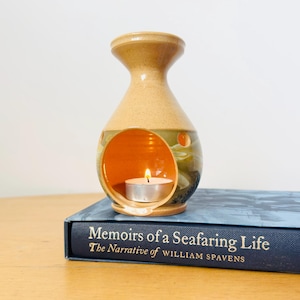 May include: A tan and green ceramic oil burner with a flared top and a circular opening containing a lit tea light candle, resting on a dark blue book titled Memoirs of a Seafaring Life The Narrative of William Spavens with gold lettering.