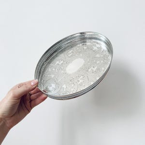 May include: A silver-coloured oval tray with a polished finish and an intricate embossed floral pattern across the base. The tray features a raised, perforated gallery rim with a delicate lattice design and a smooth, blank oval centre.