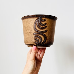 May include: A brown ceramic plant pot with a textured finish and a dark brown, stylised swirl pattern. The pot has a cylindrical shape with a slightly flared rim. The design is repeated on the front of the pot.