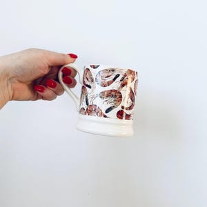 May include: White ceramic mug with a handle, featuring a repeating pattern of brown and red shrimp illustrations. The mug has a wide base and a curved handle, perfect for enjoying a warm beverage.