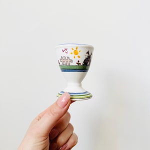 Könnte beinhalten: Ein weißer Eierbecher aus Keramik mit einem farbenfrohen handgemalten Design, das eine Sonne, einen Zaun und einen Vogel zeigt. Der Becher hat einen blau-grünen gestreiften Sockel.