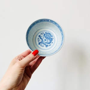 May include: A small, round, white and blue porcelain bowl. The bowl has a blue dragon design in the centre and a patterned border. The bowl is held by a hand with red nail polish against a white background.