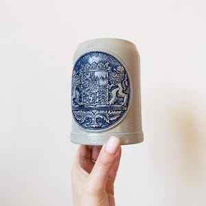 Op de afbeelding: Een lichtgrijze keramische bierpul met een blauw en wit embleem. Het embleem toont een wapen met leeuwen, een kroon en het woord "BAYERN". De pul heeft een cilindrische vorm en is ongeveer 15 cm hoog.