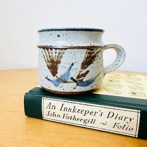 Puede incluir: Una taza de cerámica blanca con asa negra y el texto "But First, Coffee" en negrita negra. La taza se encuentra sobre un fondo borroso, sugiriendo un ambiente acogedor.