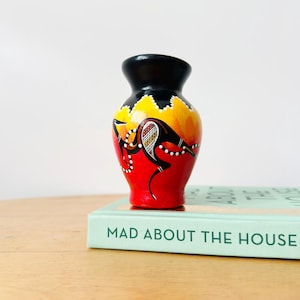 May include: A small, rounded vase with a black neck and rim, featuring a vibrant red, orange, and yellow body decorated with a stylized black kangaroo silhouette, white dot patterns, and intricate cross-hatching details on its midsection.