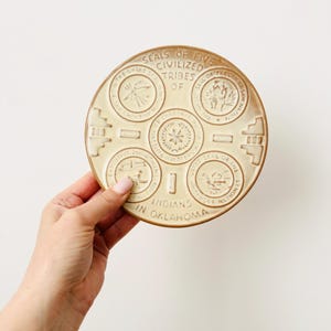 May include: A beige ceramic plate featuring embossed seals of the Five Civilized Tribes of Oklahoma. The plate has a circular design with text that reads "Seals of Five Civilized Tribes of Indians in Oklahoma."