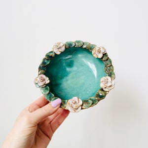 May include: A turquoise ceramic bowl with a floral design. The bowl has a glossy finish and is encircled by a border of green leaves and white and pink roses. The bowl is held by a person.