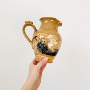May include: Tan ceramic jug with a handle and spout. The jug features a landscape design with black trees and a marbled effect. The jug is held by a person.