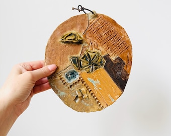 May include: A round, ceramic wall hanging with a textured surface. The artwork features a geometric design with brown, tan, and blue accents. A leather cord is attached for hanging. The piece is held by a hand.
