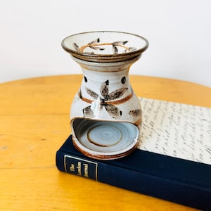 May include: A ceramic oil warmer with a speckled off-white glaze and brown painted floral motifs. The piece features a wide top basin for wax or oil and a hollow arched base for a tea light candle, resting on a dark blue book titled The London Journal.