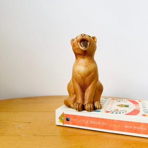 May include: A brown ceramic cat figurine with its mouth open, sitting on a book titled "The Little Book of Lykke" by Meik Wiking.
